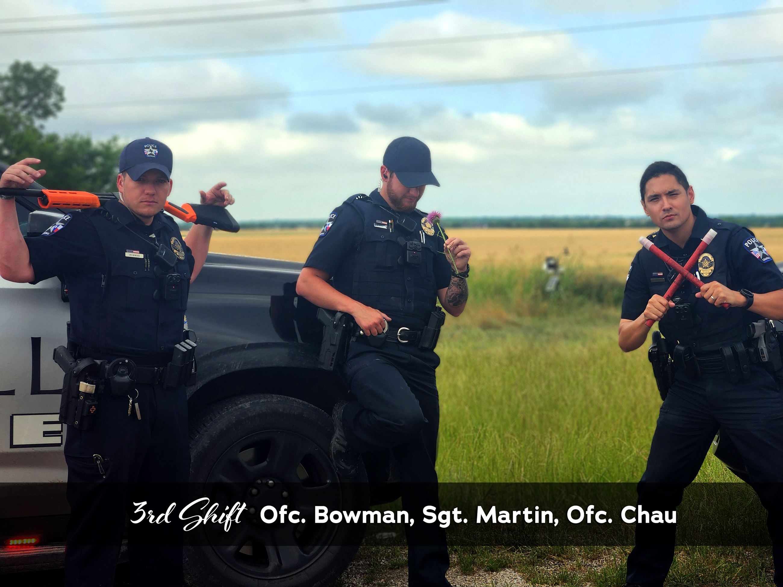 3rd patrol shift standing in front of field with squad car posing with a gun, flower, and sabors